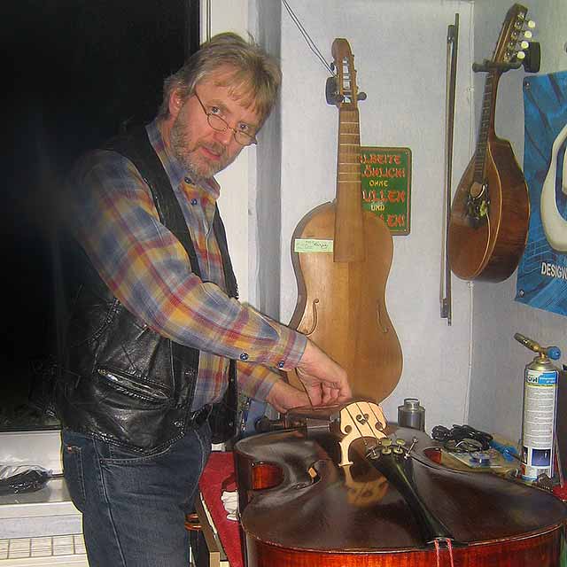 Auf dem Werktisch liegt ein repariertes Cello, an der Wand hinten hängt eine altdeutsche Gambe (Restauration) und rechts eine portugiesische Flachmandoline